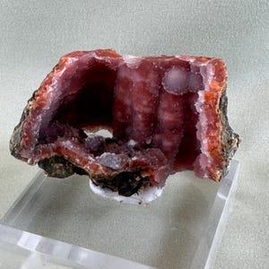 Puede incluir: Una secci&oacute;n transversal de una geoda, que muestra una vibrante exhibici&oacute;n de colores naturales. El interior revela un rico tono rojo intenso con formaciones cristalinas, enmarcado por una capa exterior naranja y marr&oacute;n oscuro contrastante. La geoda se exhibe sobre un soporte transparente.