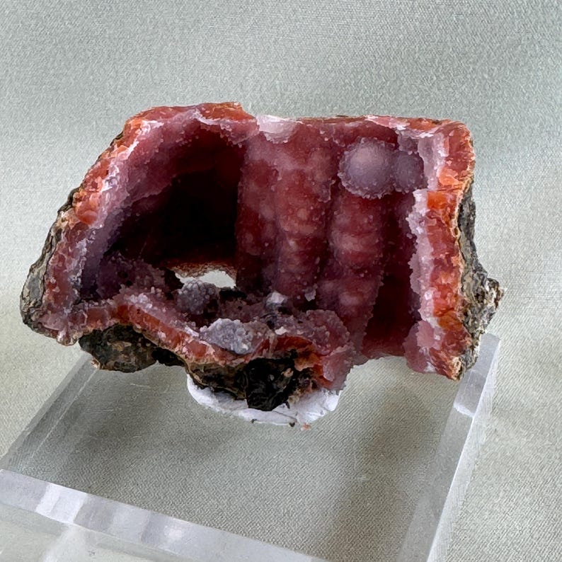 Puede incluir: Una secci&oacute;n transversal de una geoda, que muestra una vibrante exhibici&oacute;n de colores naturales. El interior revela un rico tono rojo intenso con formaciones cristalinas, enmarcado por una capa exterior naranja y marr&oacute;n oscuro contrastante. La geoda se exhibe sobre un soporte transparente.