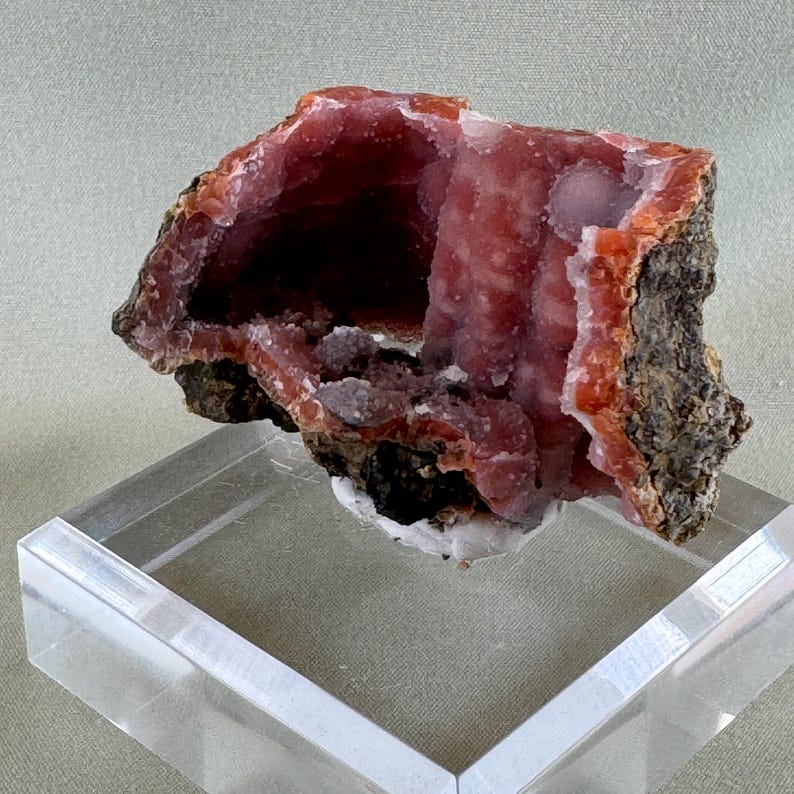 Puede incluir: Un esp&eacute;cimen mineral natural vibrante con una llamativa combinaci&oacute;n de colores y texturas. El esp&eacute;cimen presenta un interior de color rojo-rosa intenso con un exterior &aacute;spero de color marr&oacute;n oscuro. El mineral se exhibe sobre un soporte acr&iacute;lico transparente.