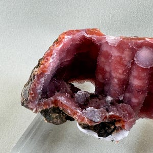 Puede incluir: Primer plano de una geoda con una cavidad, que muestra una vibrante exhibici&oacute;n de tonos rosas y rojos. La formaci&oacute;n cristalina es visible, con un exterior rugoso y texturizado y un interior liso y cristalino. La geoda est&aacute; montada sobre un soporte transparente.