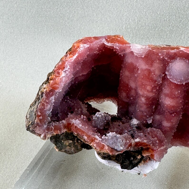 Puede incluir: Primer plano de una geoda con una cavidad, que muestra una vibrante exhibici&oacute;n de tonos rosas y rojos. La formaci&oacute;n cristalina es visible, con un exterior rugoso y texturizado y un interior liso y cristalino. La geoda est&aacute; montada sobre un soporte transparente.
