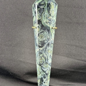 May include: A polished, six-sided crystal tower featuring a complex pattern of dark black and mottled sage green circular inclusions. The stone is held in place by two thin, gold-toned metal prongs that extend from the sides of the crystal.