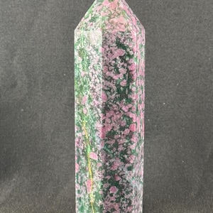 May include: A tall, faceted crystal point with a polished finish. The stone features a speckled pattern of pink and green hues, with a pointed top. The crystal is set against a dark background.