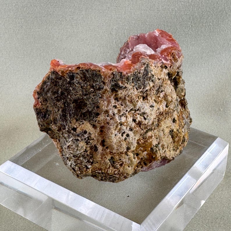 Puede incluir: Un esp&eacute;cimen mineral natural y rugoso que presenta una mezcla de texturas y colores. El exterior es marr&oacute;n y beige moteado, con una apariencia porosa. El interior revela una vibrante formaci&oacute;n de cristales rosas.