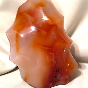 May include: A polished, freeform gemstone with a starburst shape. The stone features a blend of translucent peach, orange, and deep red hues, with some areas appearing nearly opaque. The surface has a glossy finish, and the overall form is smooth and organic.