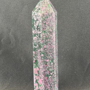 May include: A tall, polished crystal point with a flat base and a pointed top. The stone features a speckled pattern of pink and green hues. The crystal is displayed against a dark background.