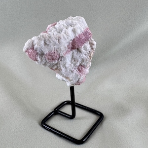 May include: A raw, natural mineral specimen featuring a white matrix with bands of pink tourmaline. The stone is mounted on a black metal display stand with a square base, showcasing the crystal's unique texture and color variations.
