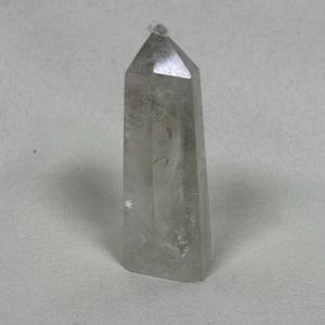 May include: A clear quartz crystal point with a faceted top. The crystal is a light gray color and has internal inclusions. The crystal is standing upright and has a rectangular base.