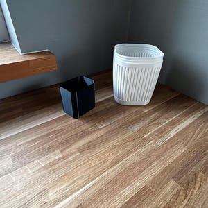 Op de afbeelding: Twee kleine containers op een oppervlak met houtnerf. Een zwarte vierkante container staat links en een witte, verticaal geribbelde container staat rechts. De achtergrond is een grijze muur.