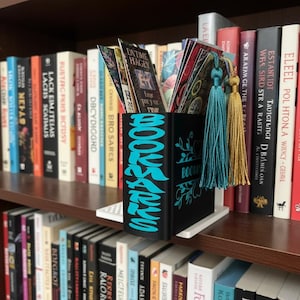 Könnte beinhalten: Ein schwarzer BOOKMARKS-Halter mit türkisfarbenen Buchstaben und floralen Akzenten, der verschiedene Lesezeichen enthält. Der Halter hat eine weiße Basis und wird in einem Bücherregal mit Büchern ausgestellt. Türkisfarbene und goldene Quasten hängen vom Halter.