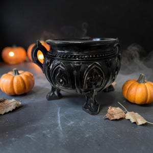 May include: A black cauldron with three legs and two handles, featuring embossed gothic arch patterns and a beaded rim. The vessel is positioned among orange pumpkins and scattered dried leaves, creating a dark, seasonal display.