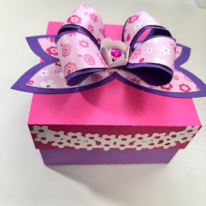 May include: A pink gift box with a large bow on top. The bow is decorated with a floral pattern in shades of pink and white, with purple accents. The box has a white floral trim.