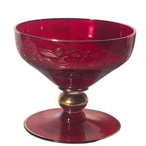 Puede incluir: Un cuenco de postre de cristal rojo rubí con borde dorado y diseño floral. El cuenco tiene una parte superior ancha y poco profunda, un tallo con un acento dorado y una base redonda con borde dorado. El cristal es transparente.