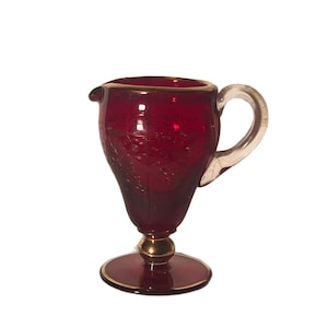 Puede incluir: Una jarra de vidrio rojo rubí con asa transparente y ribete dorado. La jarra tiene una base redonda y un diseño decorativo grabado en el vidrio. La jarra mide aproximadamente 13 cm de altura.