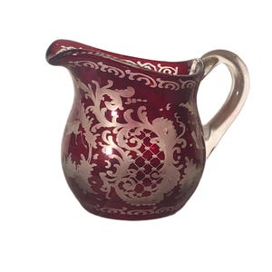 May include: A small, ruby red glass jug with a clear glass handle. The jug is decorated with a silver, ornate floral pattern. The rim of the jug has a scalloped edge.