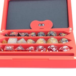 Puede incluir: Una caja de dados roja con interior negro, que contiene varios dados poliédricos de diferentes colores y diseños. Un contador rojo muestra el número 79. La caja está abierta, mostrando los dados.