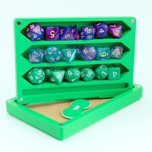 Könnte beinhalten: Eine grüne Würfelaufbewahrungsbox mit drei Würfelreihen. Die oberste Reihe enthält lila und blaue Würfel, die mittlere Reihe hat türkisfarbene Würfel und die untere Reihe grüne Würfel. Ein grüner Token befindet sich in der Box.