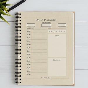 May include: A spiral-bound daily planner with a beige cover, featuring sections for schedule, date, notes, and reminders. The planner includes the text "DAILY PLANNER" and "Top a Priorliers". A black pen and a small plant are visible.