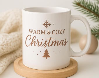 May include: White ceramic mug with the words "WARM & COZY Christmas" in brown script, along with a snowflake and Christmas tree graphic. The mug sits on a round wooden coaster. A cozy holiday theme.