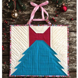 Puede incluir: Un patrón de bloque de árbol de Navidad acolchado titulado "Merry Little Bow Tree" con un lazo rojo y un árbol azul. El patrón es de Natalee Terrile y es apto para retales o FQ. El fondo es una superficie de madera con adornos navideños.
