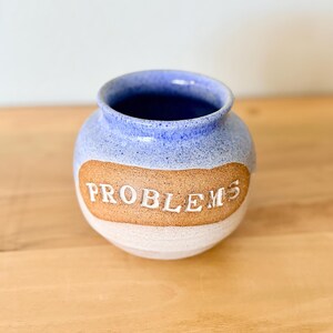 May include: A round ceramic vase with a speckled blue glaze on the top and a white base. A central band of unglazed tan clay features the word PROBLEMS embossed in white capital letters. The vase has a wide opening and a smooth, rounded body.