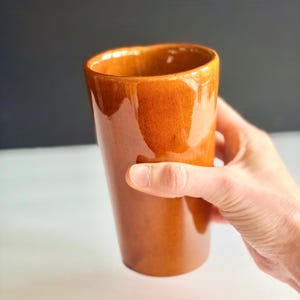 May include: A tall, glossy, brown ceramic tumbler. The cylindrical cup has a slightly flared rim and is held by a hand. The background is a gradient of black and white. The cup is likely used for drinking beverages.