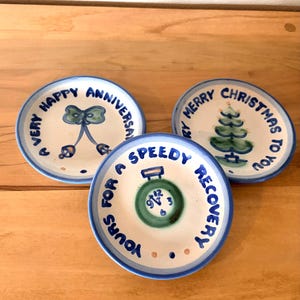 May include: Three small, round, hand-painted ceramic plates with blue rims and text. One says "Very Happy Anniversary", another "Merry Christmas to You", and the third "Yours for a Speedy Recovery". Each plate has a unique illustration.