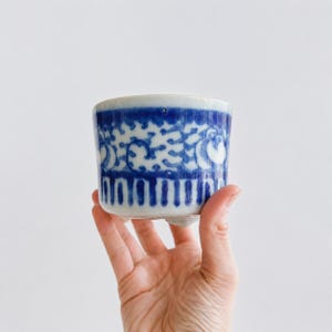 May include: A small, cylindrical ceramic cup featuring a white base with intricate blue hand-painted patterns. The design includes a band of swirling motifs above a series of vertical stripes. The cup is supported by three small, rounded feet.