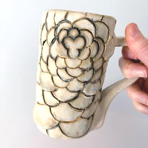 May include: A cylindrical ceramic mug with a cream-colored glaze and a hand-carved, repeating floral petal pattern. The carved lines are highlighted with a dark, charcoal-colored pigment, creating a textured, layered appearance across the surface.