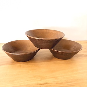 May include: Three brown wooden bowls with a textured surface are stacked on a wooden table. The bowls have a wide, shallow design with a slightly angled rim. The bowls are empty and ready to be used.