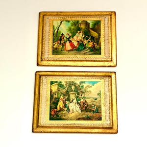 May include: Two rectangular wall art pieces featuring ornate gold frames. Each frame surrounds a central print depicting a group of figures in 18th-century attire within a lush garden setting, accented by a textured, light-colored border.