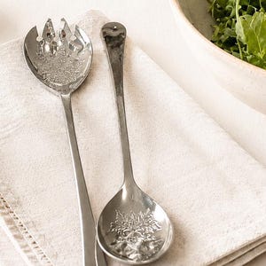 May include: A pair of silver serving utensils, a fork and a spoon, rest on a folded white linen napkin. The fork has tines and a decorative floral design. The spoon has a rounded bowl with a similar floral pattern. A bowl of greens is in the background.