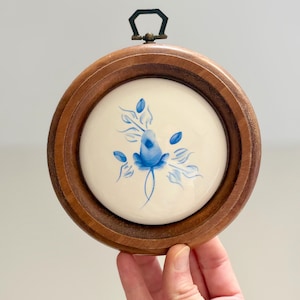 May include: A circular wall hanging featuring a white ceramic center tile with a hand-painted blue floral motif. The tile is encased in a polished dark brown wooden frame with a small metal hanging loop attached to the top center of the frame.