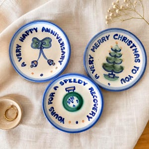May include: Three round ceramic plates with blue rims and hand-painted designs. One plate features a green bow and the text A VERY HAPPY ANNIVERSARY. Another shows a green Christmas tree and A VERY MERRY CHRISTMAS TO YOU. One shows a clock and YOURS FOR A SPEEDY RECOVERY.