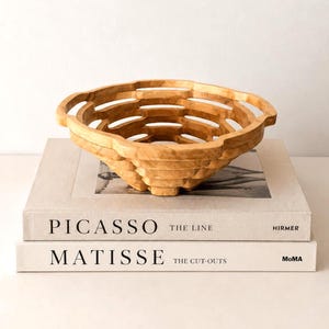 May include: A light brown wooden bowl with a lattice-like, open-weave structure and two integrated handles, resting on two stacked hardcover books titled Picasso The Line and Matisse The Cut-Outs, featuring visible wood grain patterns.
