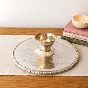 May include: A silver-toned metal serving tray with a central pedestal bowl. The tray features an intricate embossed floral pattern across its surface and a decorative rope-style edge. The bowl is smooth and polished, centered on the tray.