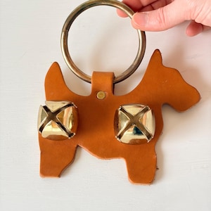 May include: A tan leather cutout in the shape of a dog featuring two large, polished gold-tone jingle bells attached to the front. A circular, dark metallic ring is secured to the top of the leather dog shape with a small metal rivet.