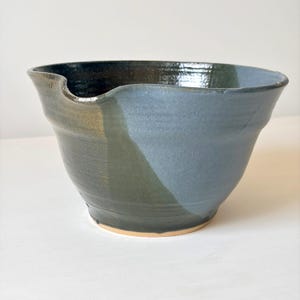 May include: A handmade ceramic mixing bowl with a wide, flared rim and a distinct pouring spout. The exterior features a two-tone glaze, with one side in a deep, glossy forest green and the other in a matte, muted slate blue, revealing a tan base.