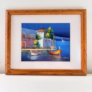 May include: A framed oil painting depicting a coastal scene with white buildings, green trees, and two boats floating on deep blue water. The artwork features a vibrant color palette and is enclosed in a light brown wooden frame with a white mat.