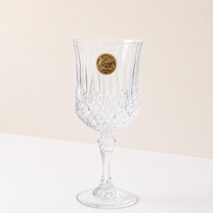 May include: A clear crystal wine glass with a detailed, cut-glass design. The glass has a round base, a slender stem, and a tulip-shaped bowl. A gold sticker with text is affixed to the glass. The glass is likely used for serving wine or other beverages.