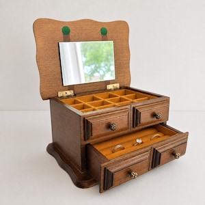 May include: A wooden jewelry box with a hinged lid featuring an interior mirror and two green circular accents. The box includes a top compartment with multiple dividers and two pull-out drawers, all lined with a mustard yellow fabric interior.