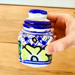 May include: A small, hand-painted ceramic jar with a blue and white striped lid and a floral design. The jar features a vibrant color palette of blue, green, yellow, and white. The jar is held in a hand.