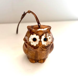 May include: A brown ceramic owl figurine with a glossy finish, featuring two large white flower-shaped eye patches with dark centers and a small orange beak. A thin brown leather strap is attached to the top of the owl for hanging purposes.