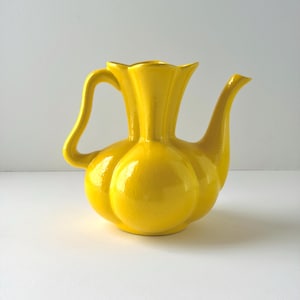May include: A bright yellow ceramic pitcher with a bulbous, segmented body, a flared scalloped rim, and a curved handle. The vessel features a smooth, glossy finish and a long, slender spout extending from the side of the rounded base.