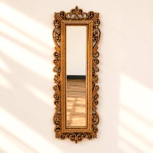 May include: A long, rectangular gold-colored mirror with an ornate, detailed frame. The frame features scrolling designs and a decorative top and bottom. The mirror is mounted on a white wall.