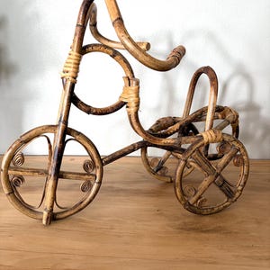 May include: A decorative bamboo tricycle wine bottle holder. The holder is made of natural bamboo with a light brown color and woven details. The tricycle has three wheels and a curved handlebar design.