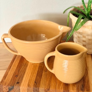 May include: Two tan ceramic kitchenware items: a large mixing bowl with a handle and spout, and a small pitcher. Both have a ribbed design. They sit on a wooden cutting board, with a potted plant in the background.