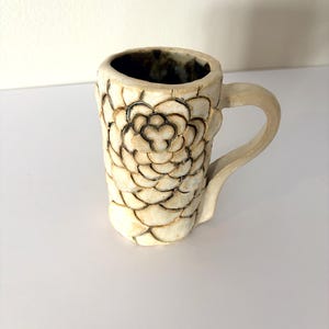 May include: Cream-colored ceramic mug with a textured, floral-like pattern. The mug has a rounded handle and a dark interior. The design features a repeating petal-like motif, creating a tactile surface. A unique, handcrafted piece for enjoying beverages.