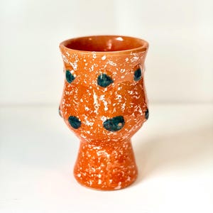 May include: A handmade ceramic vase with a speckled orange glaze and dark teal polka dots. The vase has a wide top, a bulbous midsection, and a pedestal base. The white speckles add texture and visual interest to the piece.