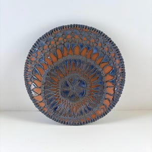 May include: A round ceramic plate featuring a terracotta-colored base with a dark blue, textured glaze pattern. The design consists of a central flower motif surrounded by concentric rings of repeating petal-like shapes and a scalloped edge.